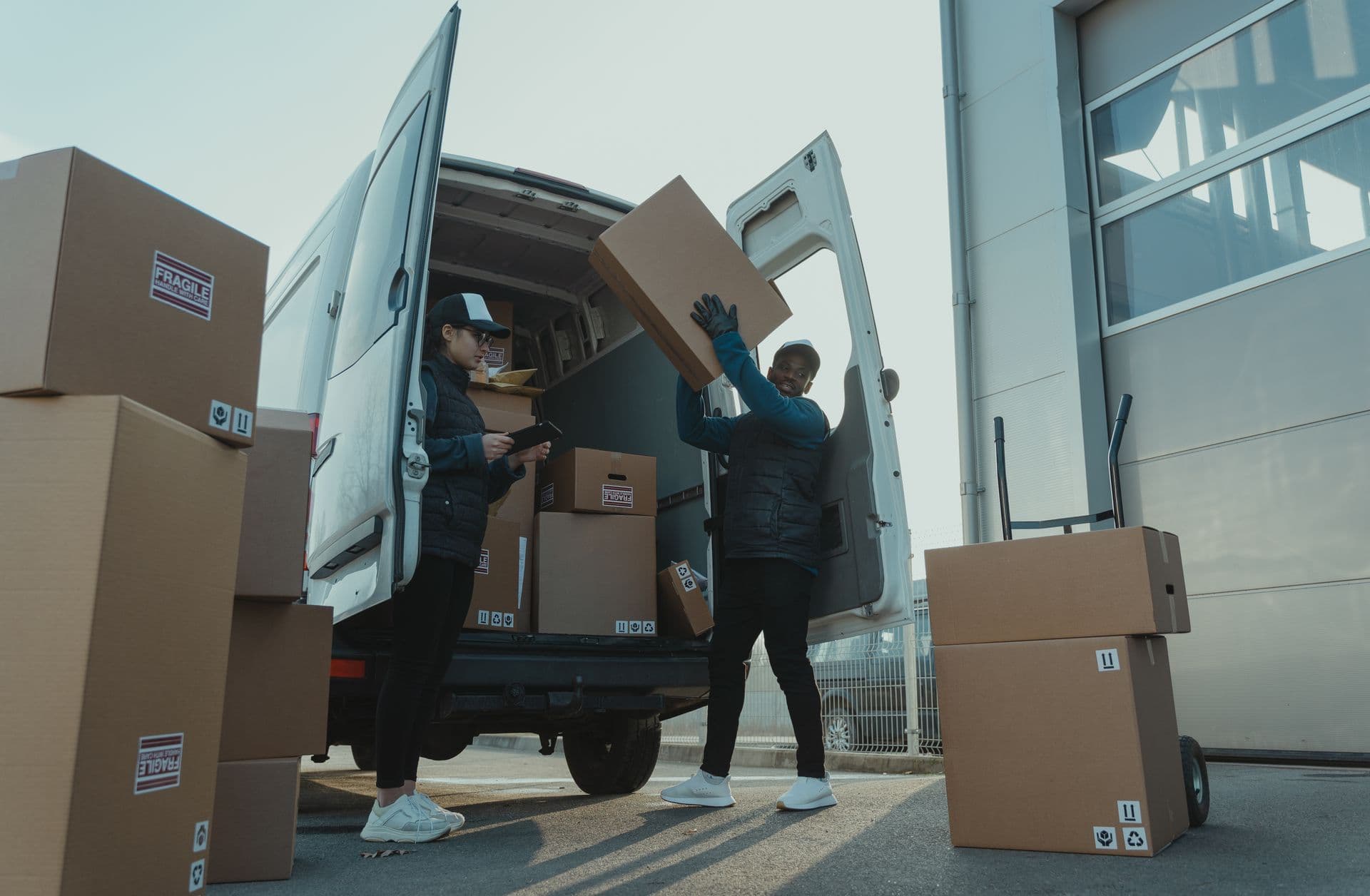 Two couriers unloading labeled cardboard boxes from a white van at a loading dock